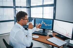 Holds cup of drink. Analysing information. Young businessman in formal clothes is in office with multiple screens. Conception of exchange and money photo