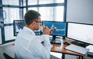 Holds cup of drink. Analysing information. Young businessman in formal clothes is in office with multiple screens. Conception of exchange and money photo