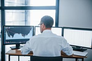 Busy employee. View from the back. Young businessman in formal clothes is in office with multiple screens. Conception of exchange and money photo