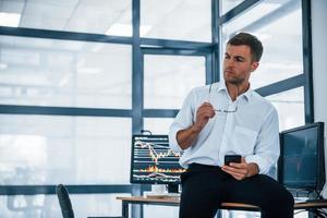 Sits on the table and having a break. With smartphone in hands. Young businessman in formal clothes is in office with multiple screens photo
