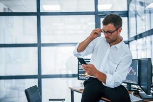Sits on the table and having a break. With smartphone in hands. Young businessman in formal clothes is in office with multiple screens photo