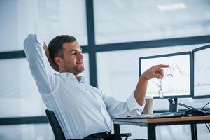 Feeling good. Conception of success. Young businessman in formal clothes is in office with multiple screens. Conception of exchange and money photo