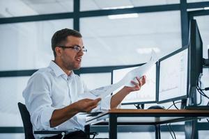 Angry employee throws paper. Conception of failure. Young businessman in formal clothes is in office with multiple screens photo