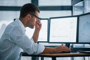 Side view. Young businessman in formal clothes is in office with multiple screens. Conception of exchange and money photo