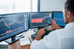 Young businessman in formal clothes is in office with multiple screens. Conception of exchange and money photo