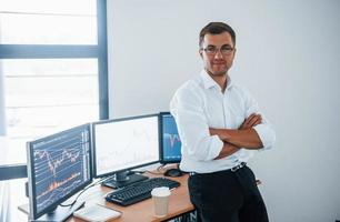 Happy employee. Young businessman in formal clothes is in office with multiple screens. Conception of exchange and money photo