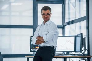 Happy employee. Young businessman in formal clothes is in office with multiple screens. Conception of exchange and money photo