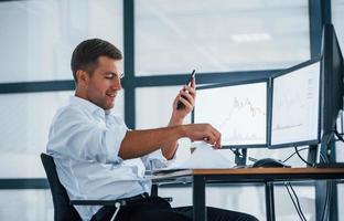Young businessman in formal clothes is in office with multiple screens. Conception of exchange and money photo