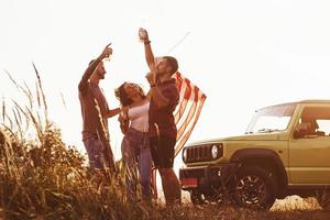 dando aplausos. los amigos tienen un buen fin de semana al aire libre cerca de su auto verde con bandera de estados unidos foto