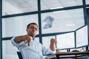 Angry employee throws paper. Conception of failure. Young businessman in formal clothes is in office with multiple screens photo