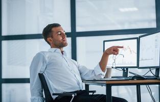 Feeling good. Conception of success. Young businessman in formal clothes is in office with multiple screens. Conception of exchange and money photo