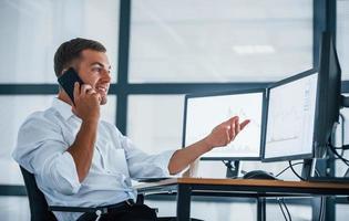 Having conversation by the phone. Young businessman in formal clothes is in office with multiple screens. Conception of exchange and money photo