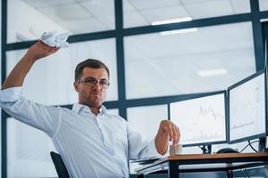 Angry employee throws paper. Conception of failure. Young businessman in formal clothes is in office with multiple screens photo
