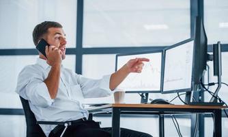 Having conversation by the phone. Young businessman in formal clothes is in office with multiple screens. Conception of exchange and money photo