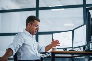Angry employee throws paper. Conception of failure. Young businessman in formal clothes is in office with multiple screens photo