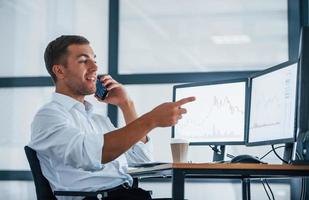 Having conversation by the phone. Young businessman in formal clothes is in office with multiple screens. Conception of exchange and money photo