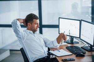 Feeling good. Conception of success. Young businessman in formal clothes is in office with multiple screens. Conception of exchange and money photo
