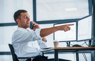 Having conversation by the phone. Young businessman in formal clothes is in office with multiple screens. Conception of exchange and money photo