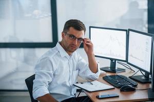 Looks at the camera. Young businessman in formal clothes is in office with multiple screens. Conception of exchange and money photo