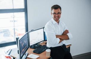 Happy employee. Young businessman in formal clothes is in office with multiple screens. Conception of exchange and money photo