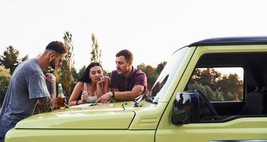 gente leyendo el mapa que yacía en el capó del automóvil. grupo de amigos alegres que tengan un buen fin de semana en un día soleado cerca de su auto verde al aire libre foto