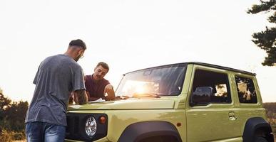 People reading map that lying on the hood of the automobile. Group of cheerful friends have nice weekend at sunny day near theirs green car outdoors photo