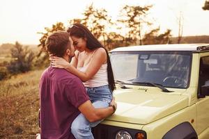 encantadora pareja besándose al aire libre. la chica se sienta en un jeep verde. hermosa tarde soleada foto