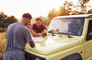 People reading map that lying on the hood of the automobile. Group of cheerful friends have nice weekend at sunny day near theirs green car outdoors photo