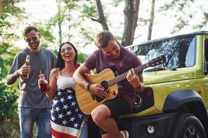 el músico toca una canción en la guitarra. los amigos tienen un buen fin de semana al aire libre cerca de su auto verde con bandera de estados unidos foto