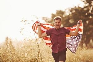 el hombre adulto corre con la bandera americana en las manos al aire libre en el campo. siente libertad en el día soleado foto