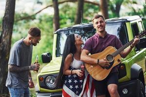 el músico toca una canción en la guitarra. los amigos tienen un buen fin de semana al aire libre cerca de su auto verde con bandera de estados unidos foto