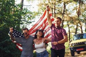 en el bosque. los amigos tienen un buen fin de semana al aire libre cerca de su auto verde con bandera de estados unidos foto