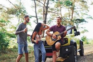 Singing songs together. Friends have nice weekend outdoors near theirs green car with acoustic guitar photo