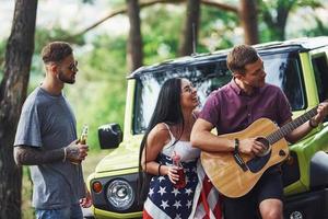 el músico toca una canción en la guitarra. los amigos tienen un buen fin de semana al aire libre cerca de su auto verde con bandera de estados unidos foto