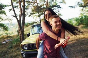 sonriendo y divirtiéndose. un par de jóvenes con alcohol se divierten en el bosque. jeep verde detrás foto