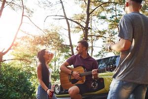 Singing songs together. Friends have nice weekend outdoors near theirs green car with acoustic guitar photo