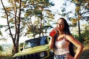 rubia con alcohol en la mano da un paseo por el bosque contra el jeep verde foto