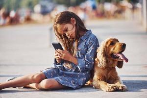 se sienta en el suelo. con teléfono inteligente en las manos. una linda niña da un paseo con su perro al aire libre en un día soleado