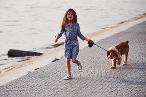 Near coastline. Cute little girl have a walk with her dog outdoors at sunny day photo