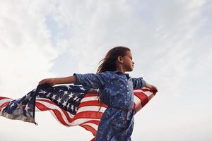 Patriotic female kid with American Flag in hands. Against cloudy sky photo