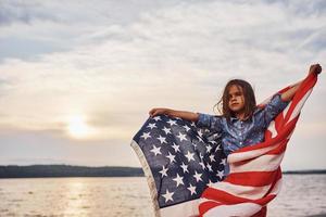 Patriotic female kid with American Flag in hands. Against cloudy sky photo