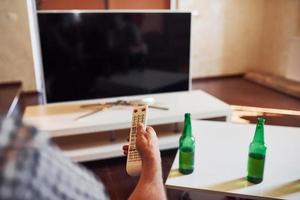 con cerveza en la mesa. vista trasera del hombre sostiene el control remoto en la mano y enciende la televisión foto