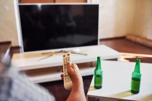 With beer on table. Backing view of man holds remote controller in hand and turning on TV