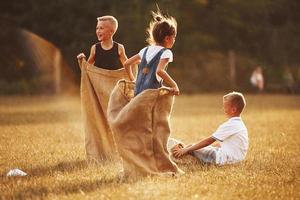 saltando carrera de sacos al aire libre en el campo. los niños se divierten durante el día soleado foto