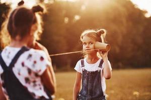 Two female kids stands in the field and talking by using string can phone photo