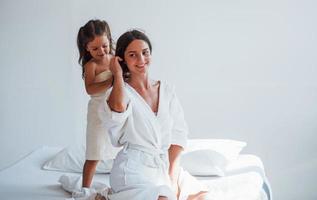 ayudando a hacer el peinado. la joven madre con su hija tiene un día de belleza en el interior de la habitación blanca foto