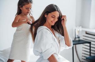Helping to made hairstyle. Young mother with her daugher have beauty day indoors in white room