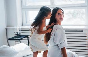 ayudando a hacer el peinado. la joven madre con su hija tiene un día de belleza en el interior de la habitación blanca foto