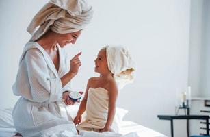 usando crema para aclarar la piel. la joven madre con su hija tiene un día de belleza en el interior de la habitación blanca foto