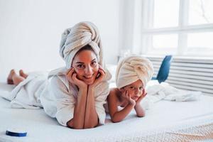 Lying down on white bed together. Young mother with her daugher have beauty day indoors in room photo